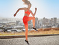 Une femme en top de sport orange et leggings saute sur un trottoir avec vue sur la ville.