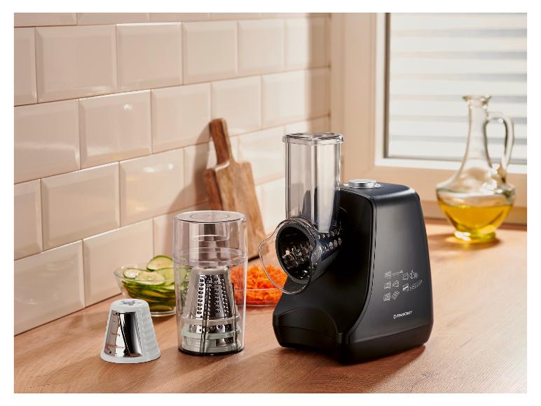 Robot culinaire noir avec accessoires, légumes râpés et huile dans une cuisine.