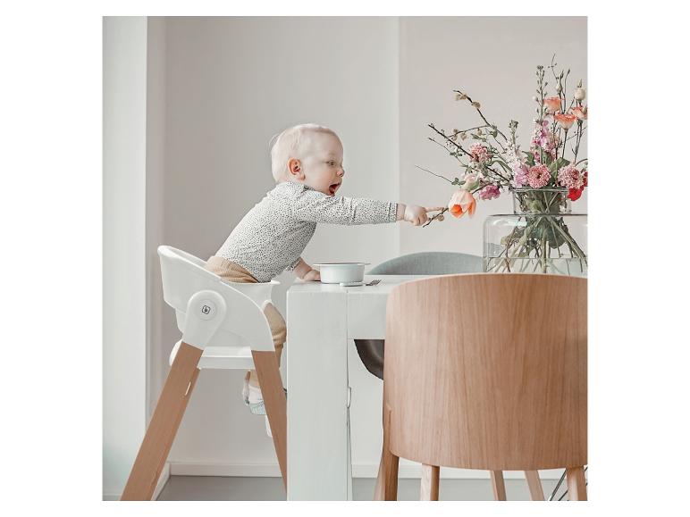 Bébé dans une chaise haute blanche tendant la main vers les fleurs sur la table.