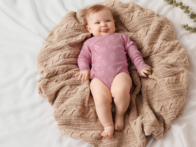 Un bébé dans un body rose à imprimé moutons est allongé sur une couverture en tricot beige.