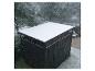 Abri de jardin noir avec de la neige sur le toit, avec des arbres et une pelouse enneigés en arrière-plan.