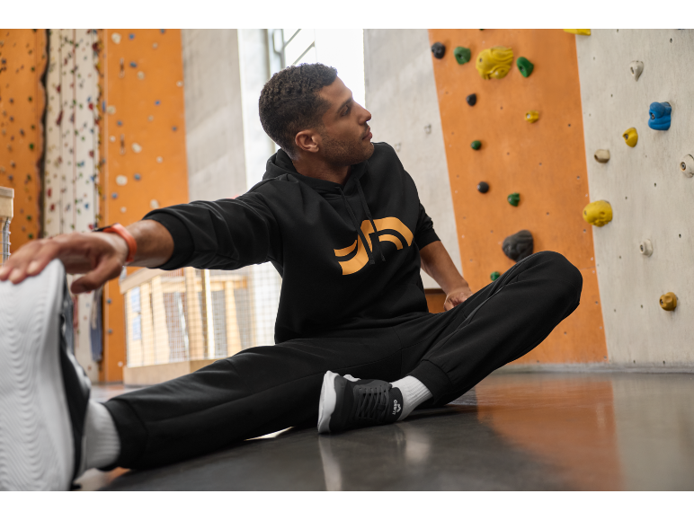 Homme en sweat à capuche et pantalon de survêtement noirs s'étirant sur le sol devant un mur d'escalade.