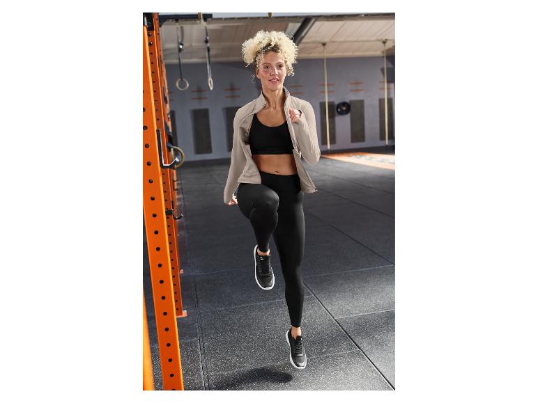 Femme en tenue de sport noire et veste beige, s'entraînant dans une salle de sport.