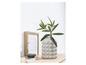 Un vase gris avec des bosses, rempli de branches vertes, à côté d'un cadre en bois.