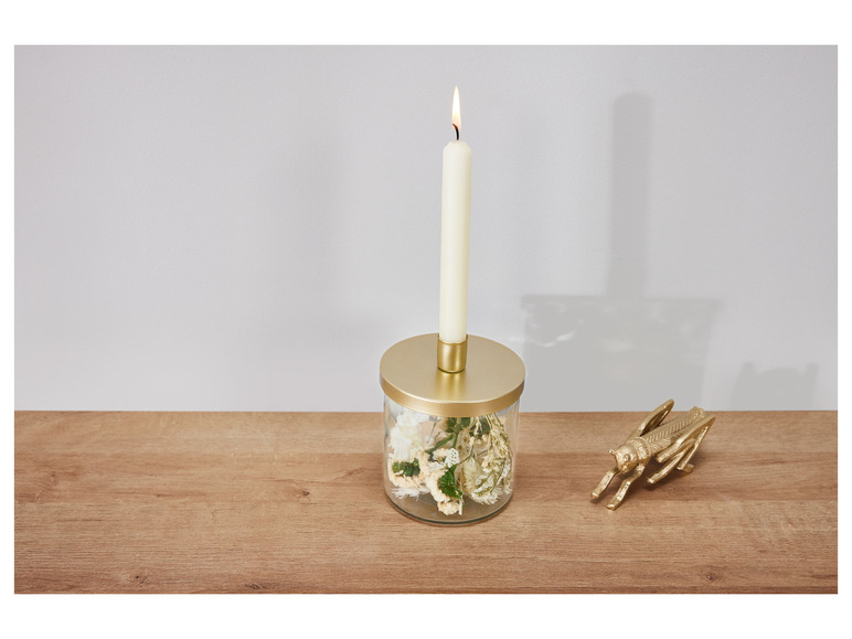 Une bougie allumée dans un bocal en verre avec des fleurs séchées et une sauterelle dorée sur une table en bois.