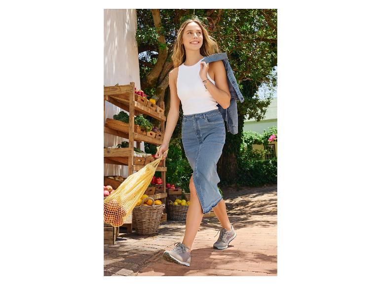 Une femme marche avec une jupe en jean et un sac en filet plein de fruits.