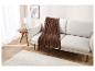 Une couverture en tricot marron avec des franges repose sur un canapé beige clair dans un salon minimaliste.