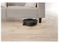 Robot aspirateur noir nettoyant sous un canapé clair sur un sol en bois.
