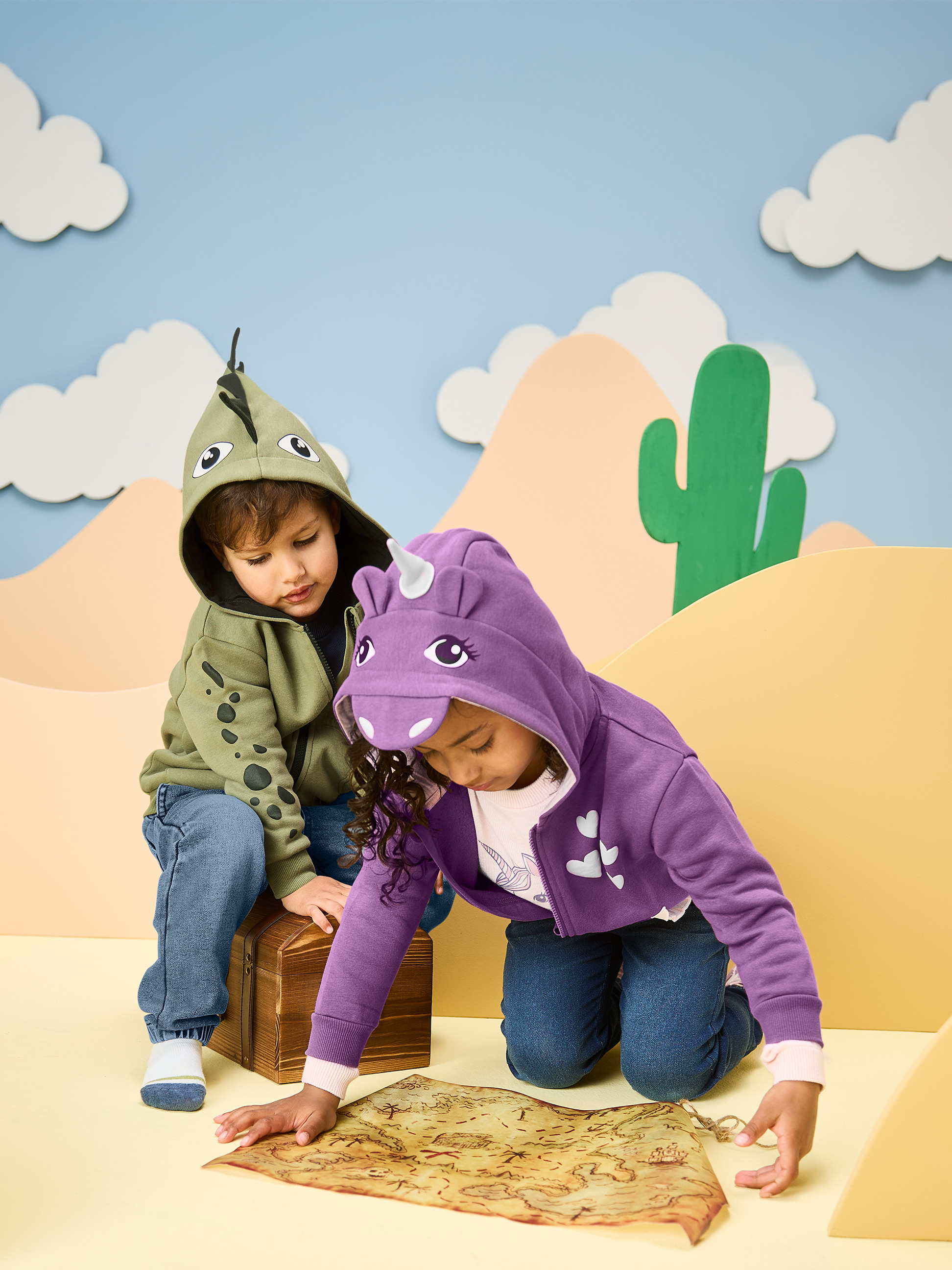 Enfants en sweats à capuche dinosaure et licorne, regardant une carte au trésor.