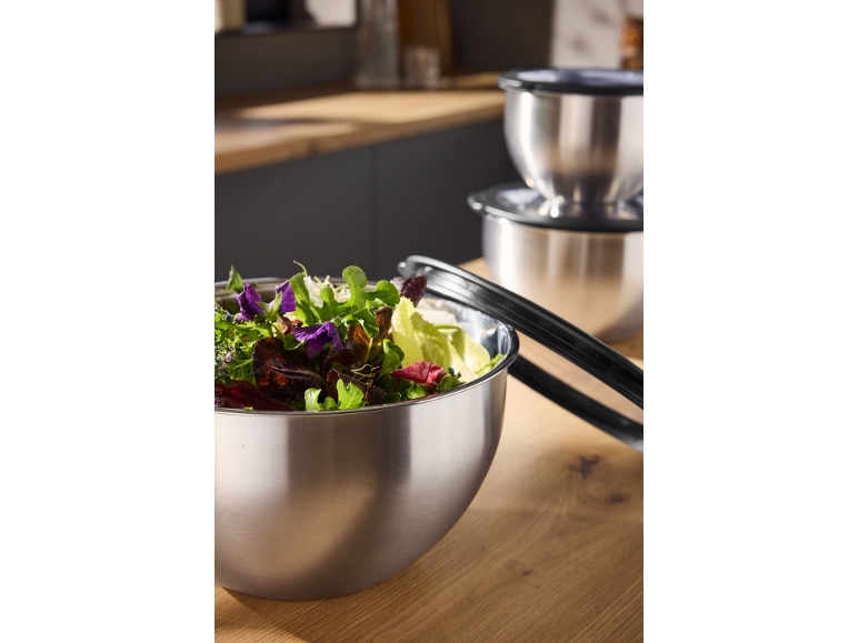 Saladiers en acier inoxydable avec salade fraîche et pinces de cuisine sur un comptoir en bois.