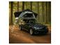 Voiture sombre avec une tente de toit et une échelle, garée sur un chemin de terre en forêt.