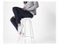 Une femme est assise sur un tabouret blanc, vêtue d'un pull en maille gris, d'un pantalon bleu foncé et de baskets blanches.
