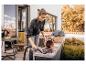 Femme aspirant terre et fleurs sur une terrasse, avec plantes et maison en arrière-plan.