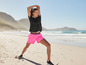 Femme en short rose et top noir faisant de l'exercice sur la plage.
