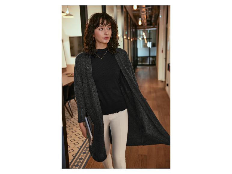 Une femme portant un long cardigan gris, un haut noir et un pantalon blanc.