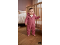 Bébé en combinaison rose à capuche debout près d'un lit en bois dans une chambre d'enfant.