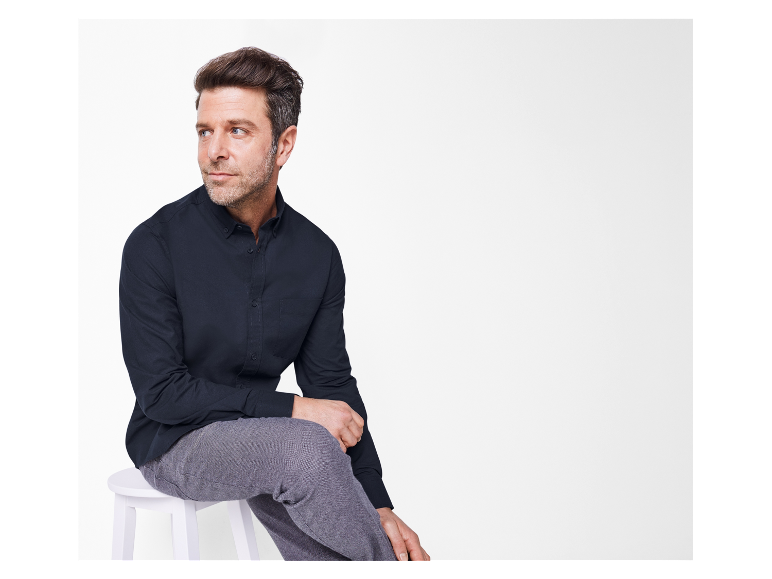 Un homme vêtu d'une chemise bleu foncé et d'un pantalon gris est assis sur un tabouret blanc.