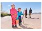 Deux enfants en vestes polaires et pantalons d'extérieur jouent sur une plage de sable, parents en arrière-plan.
