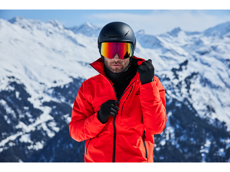 Homme avec casque de ski, lunettes et veste rouge dans les montagnes enneigées.