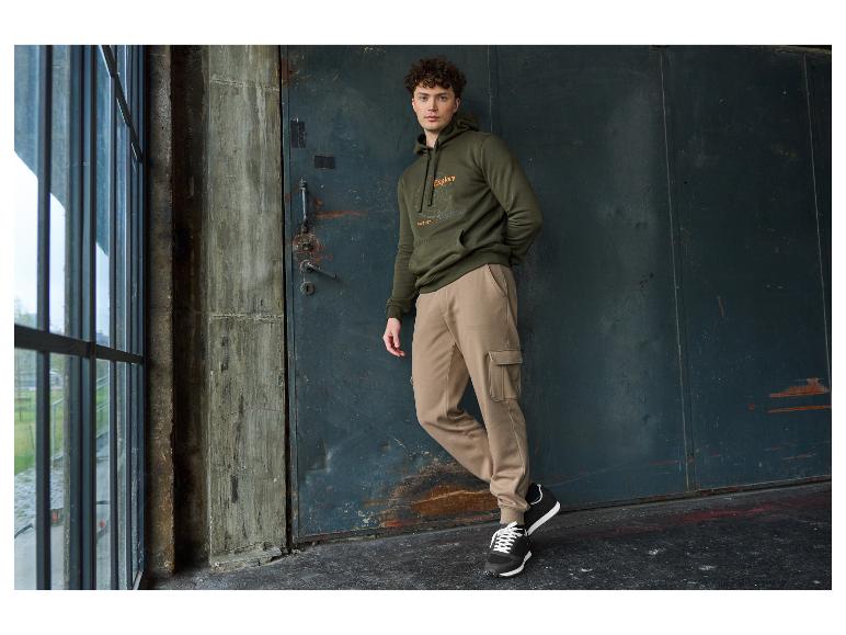 Homme en sweat à capuche vert olive et pantalon cargo beige, appuyé contre un mur.