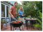 Homme grille des steaks et des tomates, femme apporte du pain dehors dans le jardin.