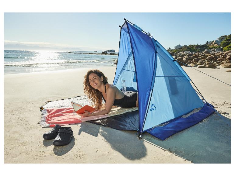 Femme lisant dans un abri de plage bleu sur une plage ensoleillée avec la mer en arrière-plan.