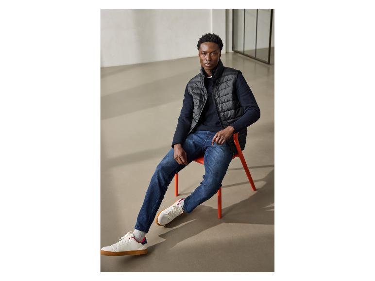 Homme en pull bleu marine, gilet noir, jean et baskets blanches, assis sur une chaise orange.