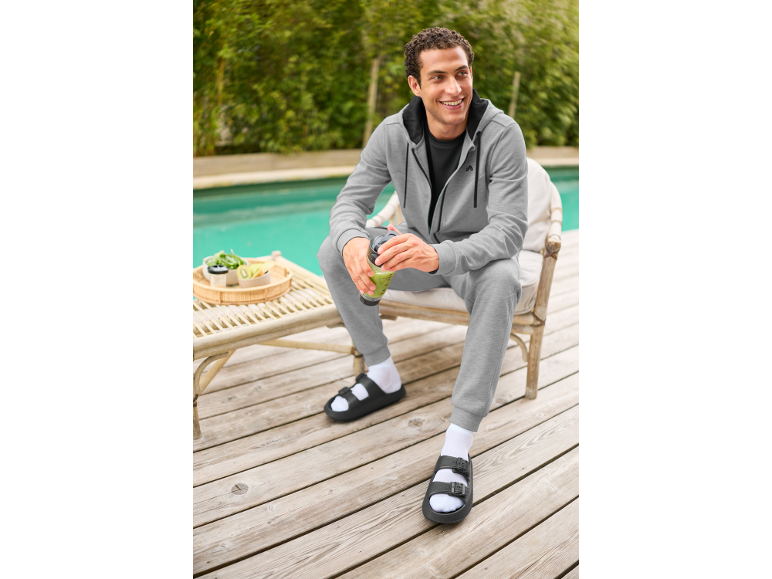 Homme en survêtement gris et sandales noires avec chaussettes blanches, souriant et tenant un smoothie vert.