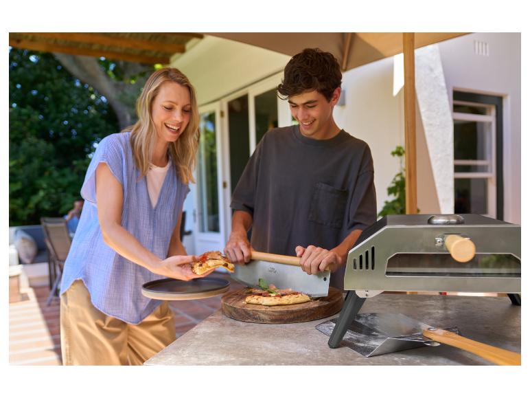 Femme et adolescent coupant une pizza en extérieur avec un four à pizza.