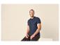 Homme souriant en polo marine et jean foncé, assis sur une surface claire.