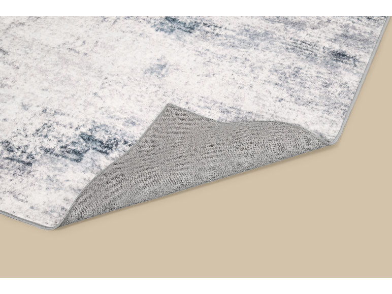 Tapis gris et blanc à motif abstrait, avec un coin replié, révélant son envers texturé.