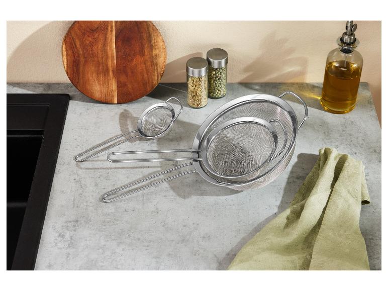 Ensemble de trois passoires en acier inoxydable de différentes tailles sur un comptoir de cuisine avec planche à découper et épices.