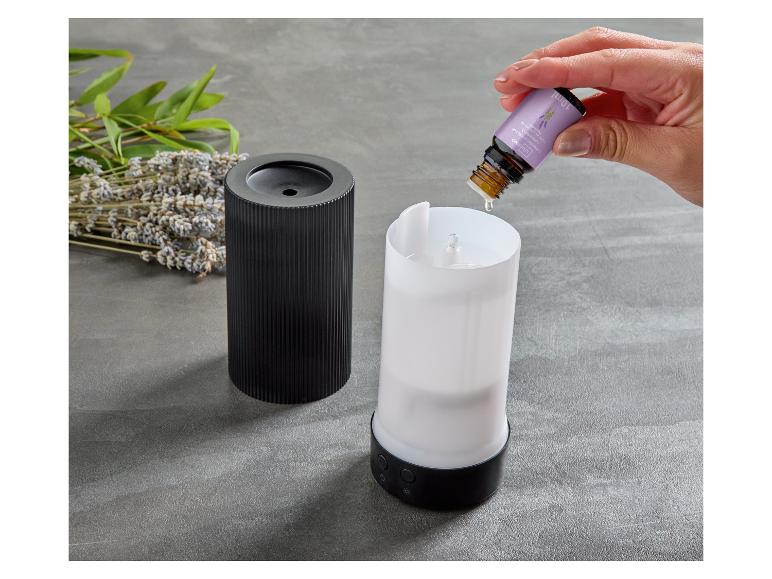 Une main ajoute de l'huile essentielle de lavande à un diffuseur d'arômes, avec de la lavande séchée et des feuilles vertes à proximité.