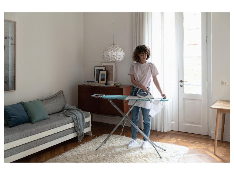 Femme repassant une chemise blanche sur une planche à repasser dans un salon avec canapé et tapis.