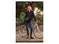 Femme en veste noire et jean, debout à côté d'un vélo avec un sac à bandoulière.