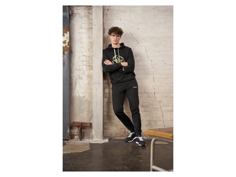 Homme en sweat à capuche et pantalon de jogging noirs Jeep, appuyé contre un mur.