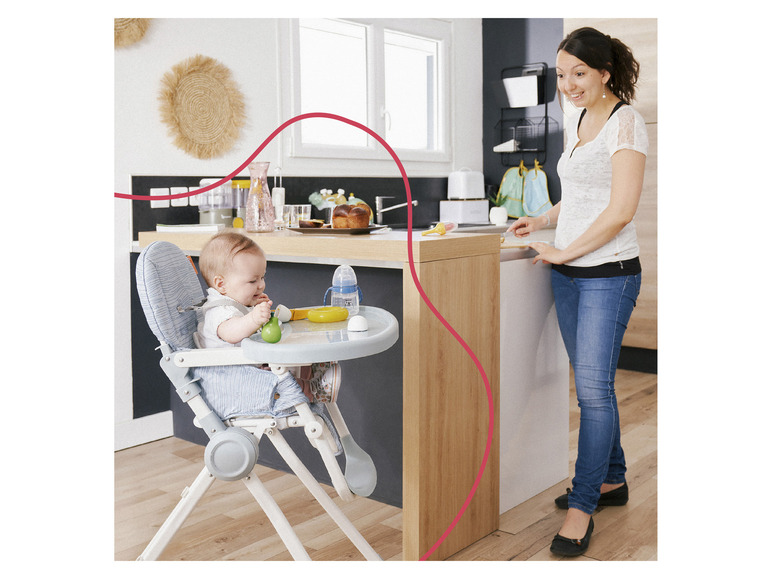 Un bébé est assis dans une chaise haute bleue et mange tandis qu'une femme est debout dans la cuisine.