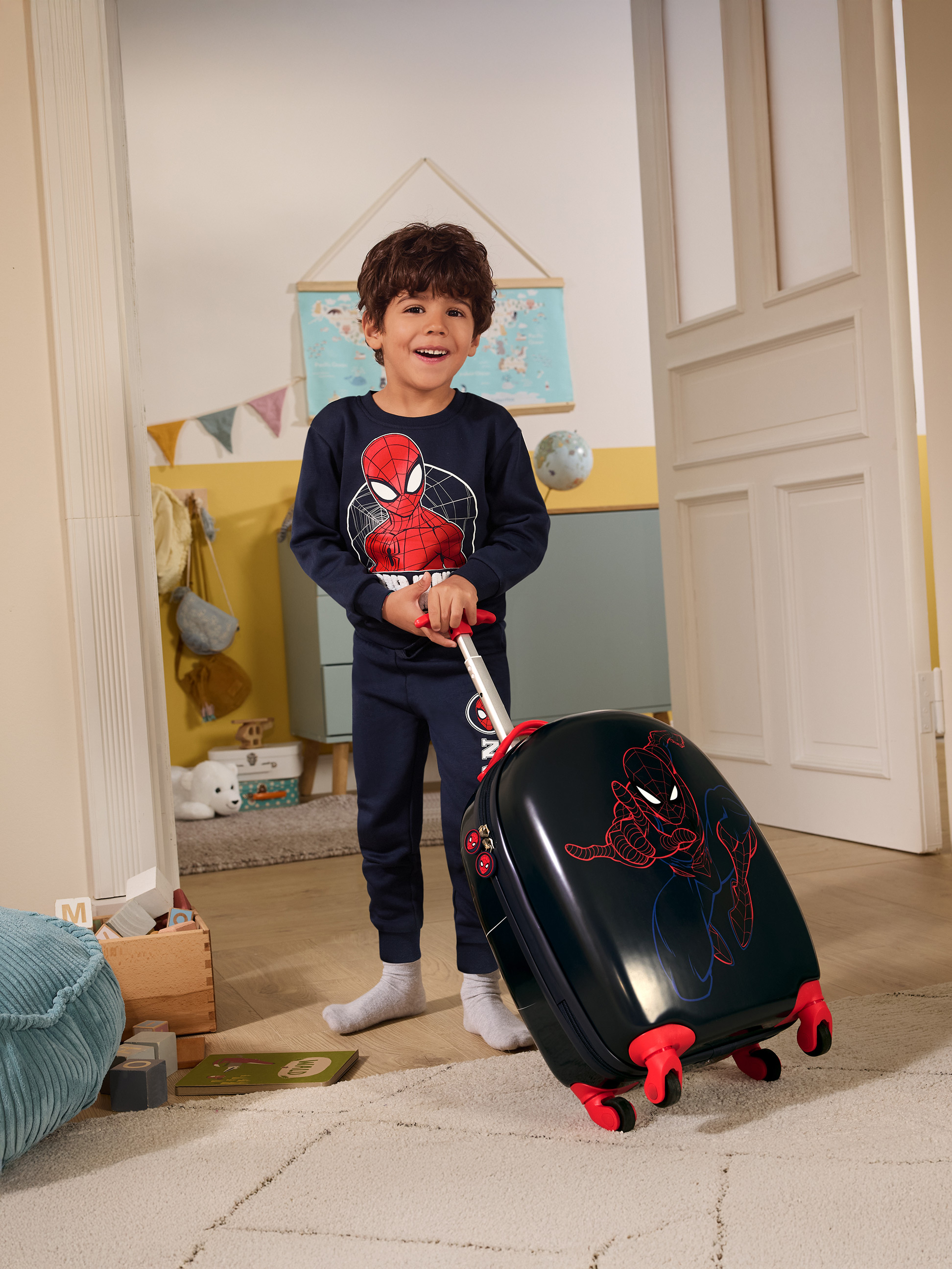 Garçon en pyjama Spiderman tirant une valise Spiderman dans une chambre d'enfant.