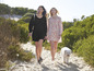 Deux femmes, l'une en robe noire, l'autre en robe à fleurs, marchent avec un chien blanc sur un chemin de sable.