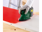 Ponceuse sans fil retirant de la peinture rouge d'une surface blanche, tenue par une main gantée.