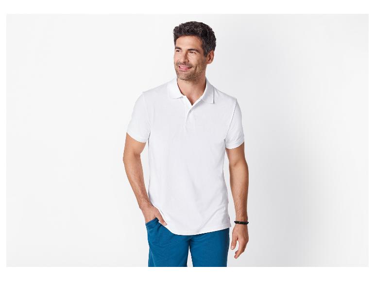 Homme en polo blanc et short bleu, souriant, la main dans la poche.