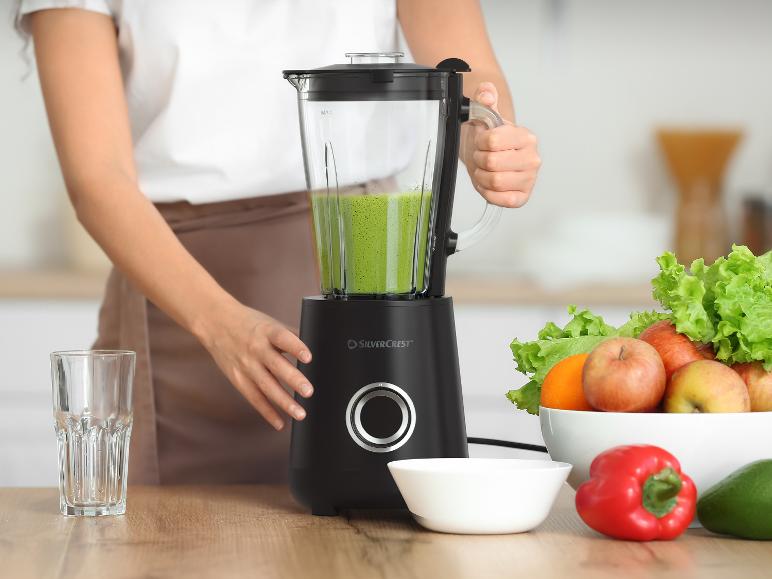 Une femme prépare un smoothie vert dans un mixeur, avec des fruits et légumes frais sur la table.