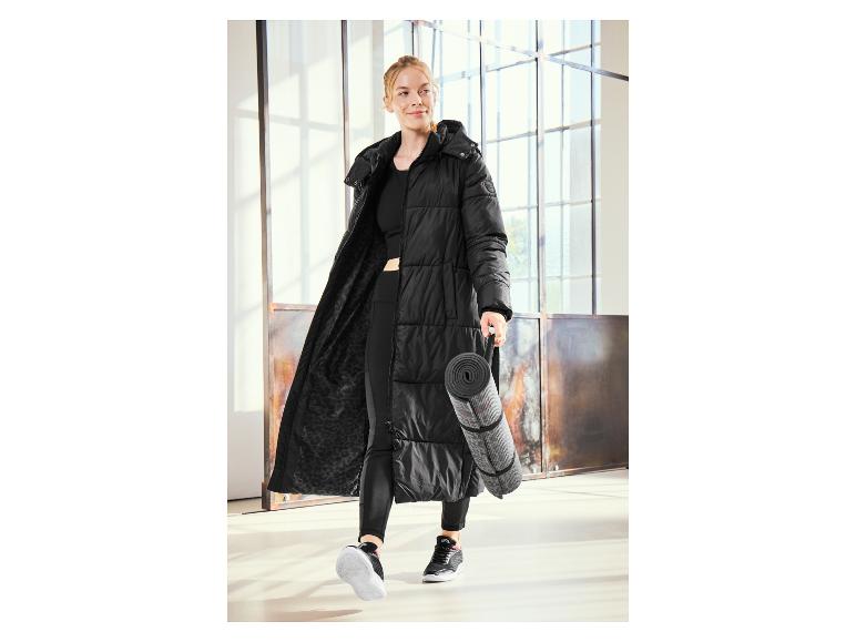 Une femme vêtue d'un long manteau noir et de leggings noirs tient un tapis de yoga.