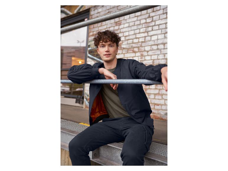 Homme en blouson aviateur noir et pantalon assis sur une structure métallique.