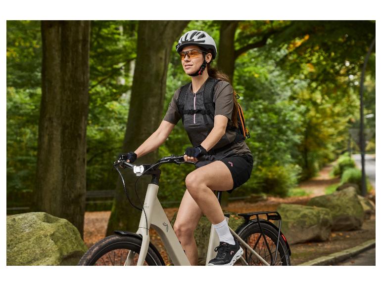 Femme sur un vélo électrique avec casque, lunettes et sac à dos sur un chemin forestier.
