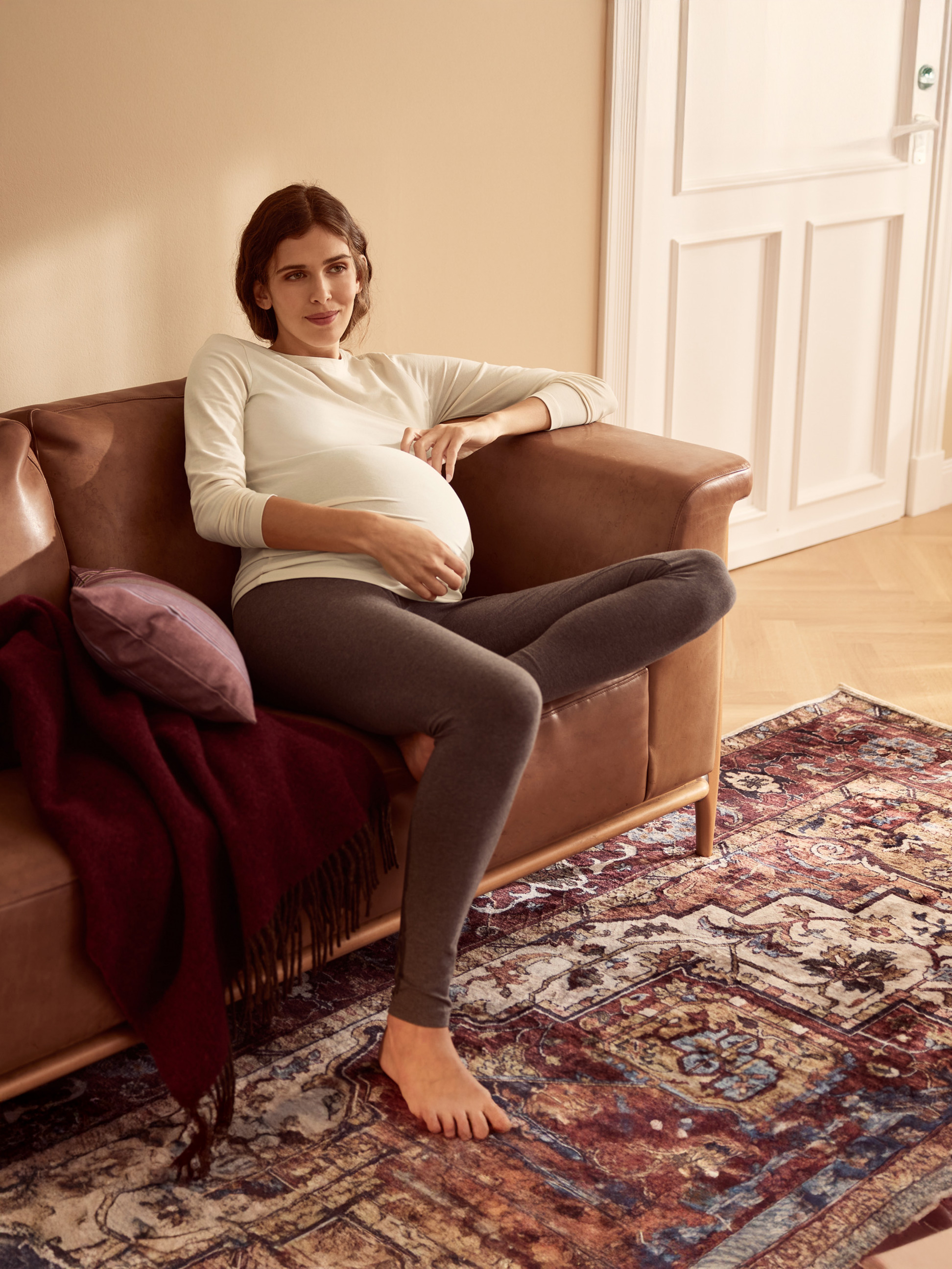 Femme enceinte en leggings et haut confortables, assise sur un canapé marron.