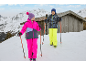 Deux enfants en tenue de ski colorée avec des bâtons de ski, marchant dans la neige près d'un chalet.