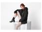 Femme rousse en doudoune noire et pantalon beige, assise sur un cube blanc.