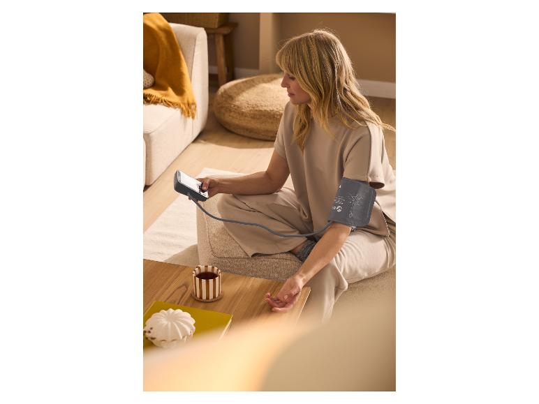 Femme mesurant sa tension artérielle avec un tensiomètre au bras, assise par terre avec une tasse de thé.