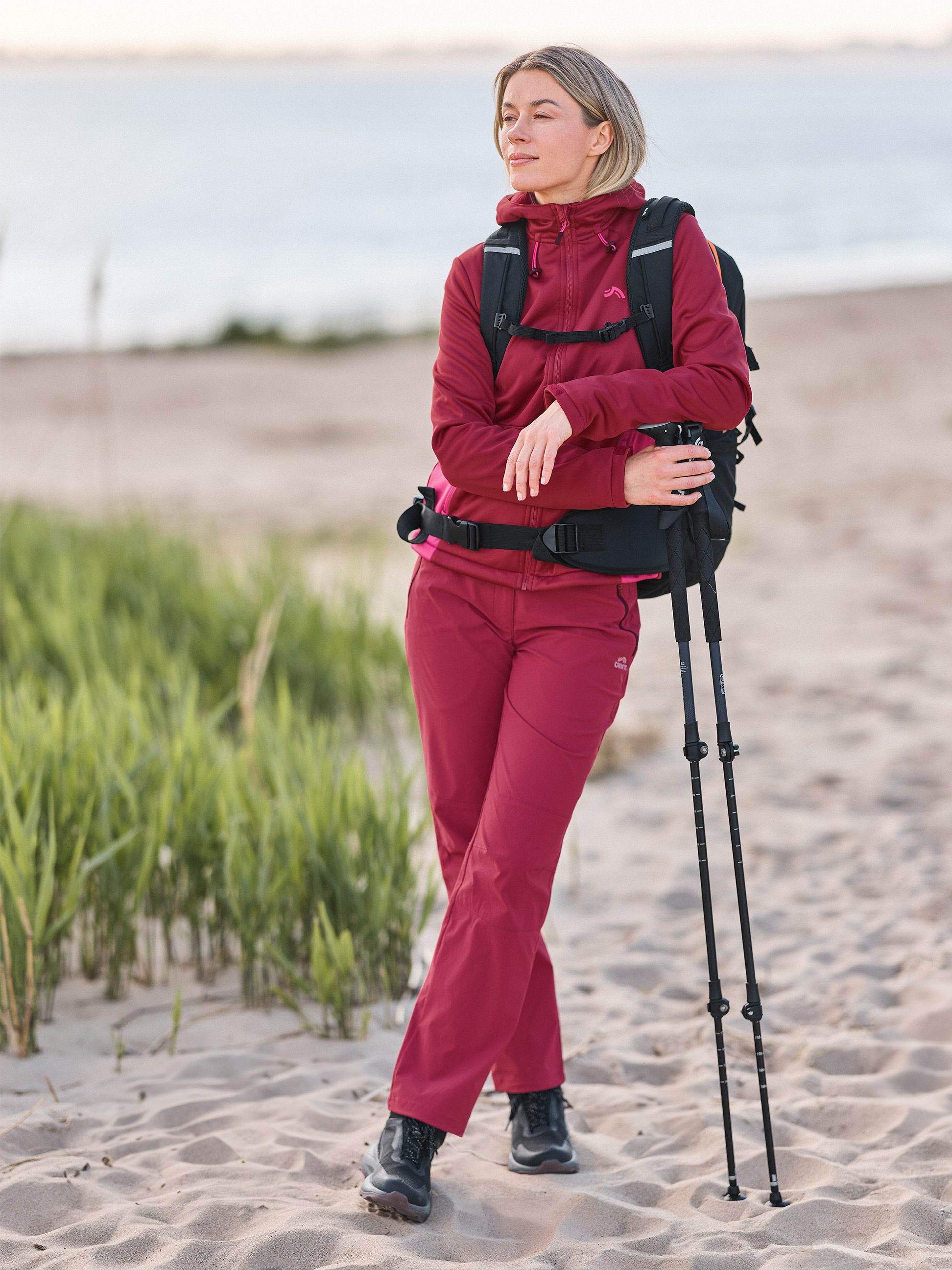 Femme en tenue de randonnée et sac à dos sur la plage, tenant des bâtons de marche.